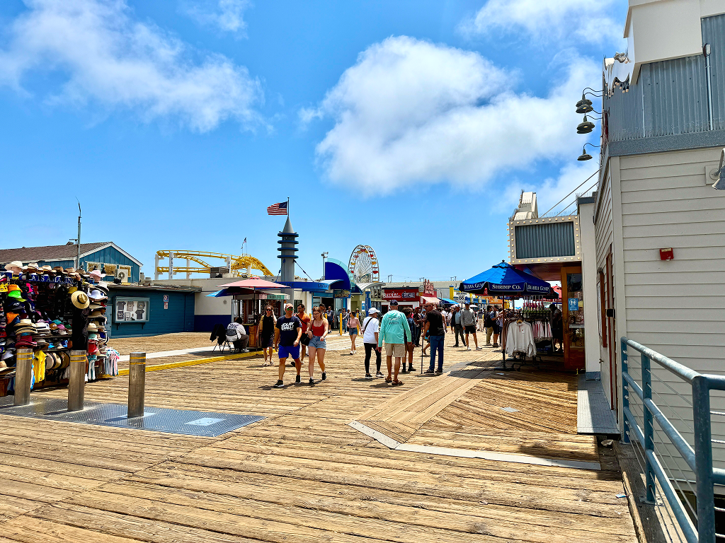 View of the Santa Monica Pier with souvenir shops on both sides and the Pacific Park amusement rides visible in the background.