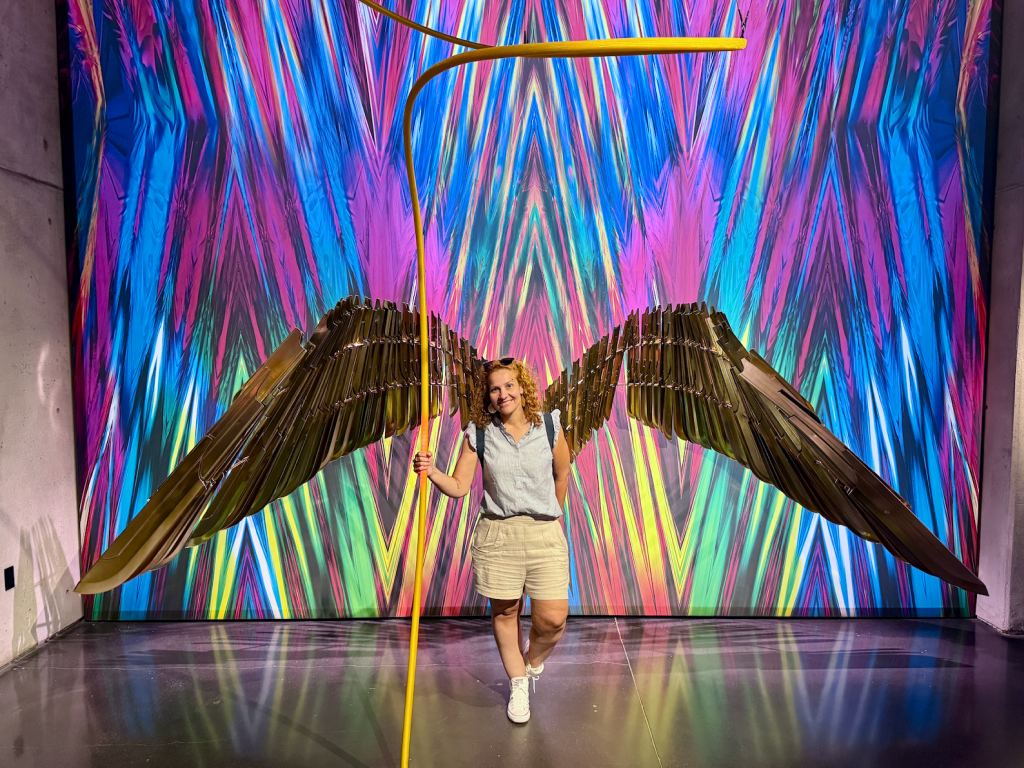 Woman holding Wonder Woman’s golden lasso with the character’s wings displayed in the background at the DC exhibit.