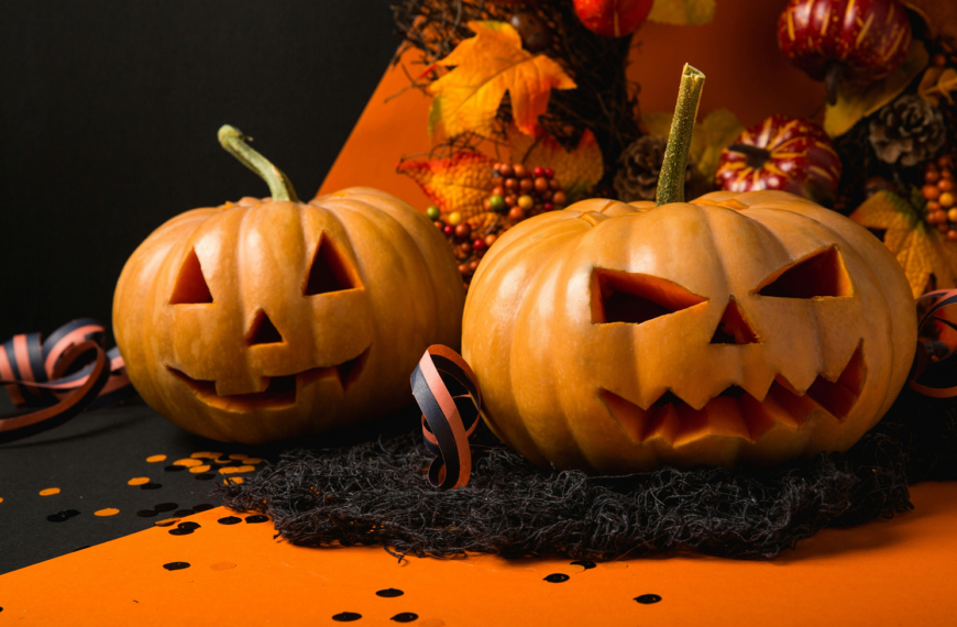 Two Halloween pumpkins surrounded by festive confetti and streamers in classic orange, black, and purple colors, creating a playful and celebratory atmosphere.