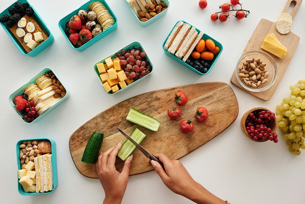 Overhead view of a person slicing cucumber on a wooden board, surrounded by bento lunchboxes filled with sandwiches, fruit, pistachios, and cheese for back to school meal prep.